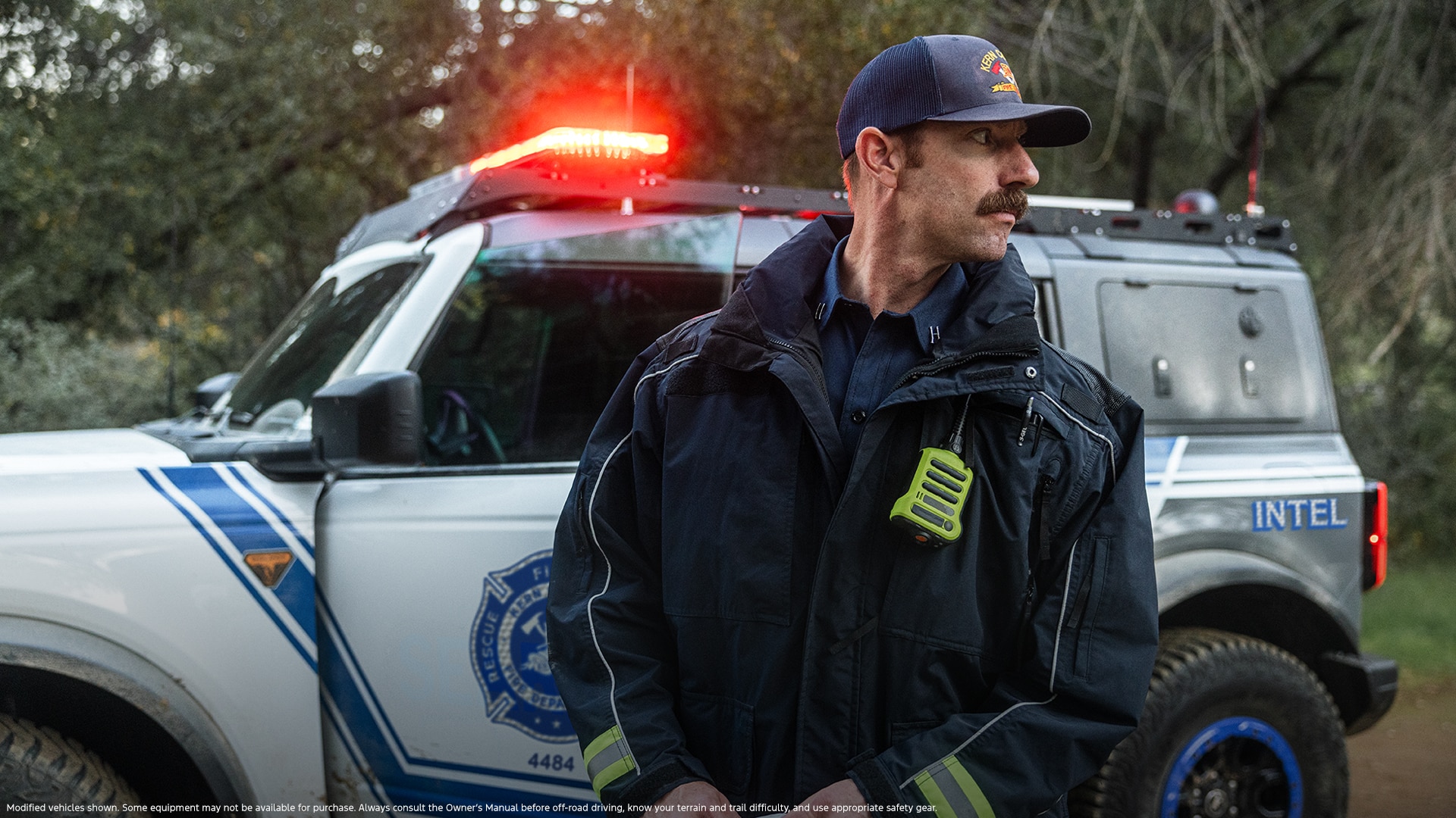 A man in uniform stands in front of a white Bronco equipped with emergency lights on top.